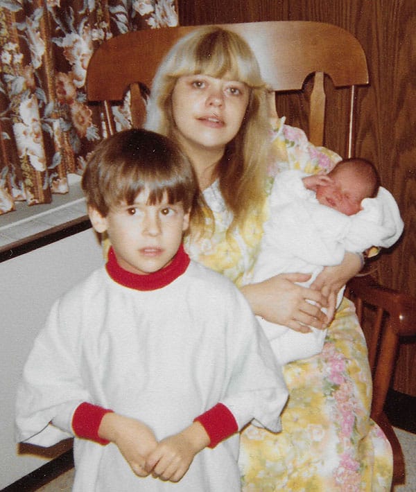 my mom sitting in a chair holding my newborn brother with me standing in front of them